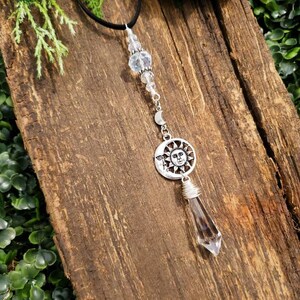 May include: A silver sun and moon charm with a clear crystal point pendant hanging from a black cord necklace. The necklace is adorned with clear crystal beads.