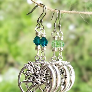 Puede incluir: Pendientes plateados de hadas y luna con cuentas de cristal verde y azul verdoso. Los pendientes cuelgan de una cuerda de hilo natural sobre un fondo verde borroso. El hada est&aacute; representada con los brazos extendidos.