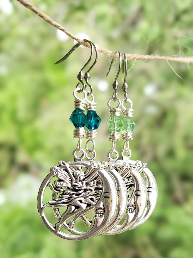 Puede incluir: Pendientes plateados de hadas y luna con cuentas de cristal verde y azul verdoso. Los pendientes cuelgan de una cuerda de hilo natural sobre un fondo verde borroso. El hada est&aacute; representada con los brazos extendidos.