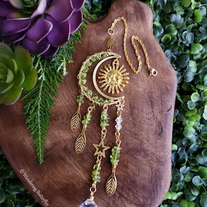 May include: A gold sun and moon dreamcatcher with green aventurine beads, gold leaves, and a clear crystal prism hanging from it. The dreamcatcher is hanging from a gold chain on a wooden surface.