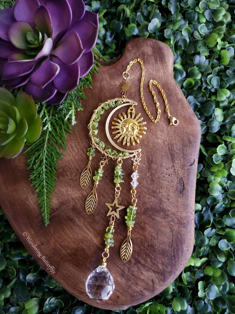 May include: A gold sun and moon dreamcatcher with green aventurine beads, gold leaves, and a clear crystal prism hanging from it. The dreamcatcher is hanging from a gold chain on a wooden surface.
