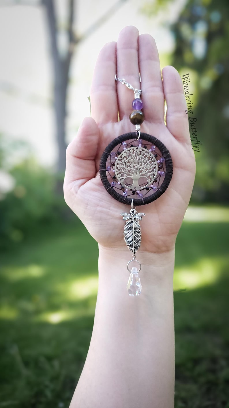 K&ouml;nnte beinhalten: Ein handgefertigter Traumf&auml;nger mit einem silbernen Lebensbaum-Design in der Mitte. Der Traumf&auml;nger hat einen dunkelbraunen gewebten Ring, verziert mit lila Perlen. Eine silberne Feder und ein klarer Kristall h&auml;ngen darunter. Der Text "Wandering Butterfly" ist sichtbar.