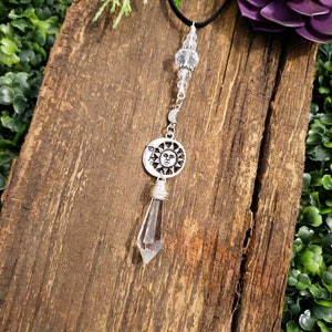 May include: A silver sun and moon charm hangs from a black cord with a clear crystal point pendant. The charm features a detailed sun face with a crescent moon.