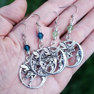 Puede incluir: Pendientes de hadas plateados con dise&ntilde;o de media luna. Cada pendiente presenta una silueta de hada dentro de la luna, acentuada con cuentas azules o verdes. Los pendientes se sostienen en una mano, mostrando sus intrincados detalles.