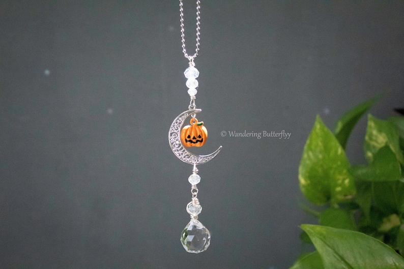 May include: A silver crescent moon charm with a small orange pumpkin hanging from it. The charm is attached to a silver chain with clear crystal beads. A large clear crystal hangs from the bottom of the charm.