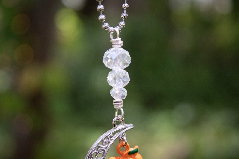 May include: A silver chain necklace with a crescent moon charm and a small orange pumpkin charm. The necklace features three clear crystal beads.