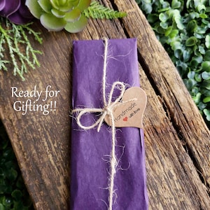 May include: A gift wrapped in deep purple paper, tied with twine, and a heart-shaped tag that reads "Handmade with love." The package rests on a rustic wooden surface, with greenery and succulents in the background. Text on the left says "Ready for Gifting!!"