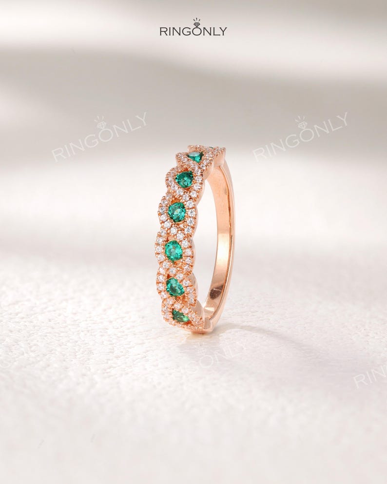 Op de afbeelding: Ros&eacute;gouden ring met een rij smaragdgroene edelstenen afgewisseld met secties van kleine, heldere edelstenen. De ring heeft een delicaat, verweven ontwerp. De merknaam "RINGONLY" is bovenaan zichtbaar.