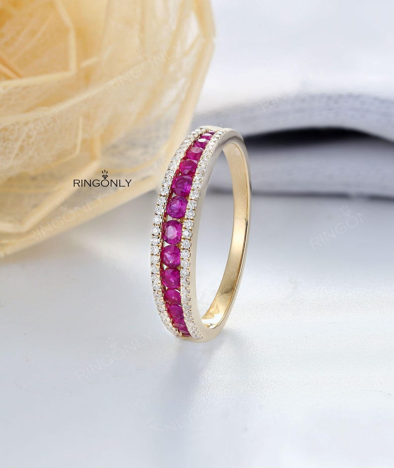 May include: A gold ring with a central row of bright red gemstones, bordered by a row of smaller, clear stones. The ring is displayed against a soft, neutral backdrop, with the brand name "RINGONLY" visible.