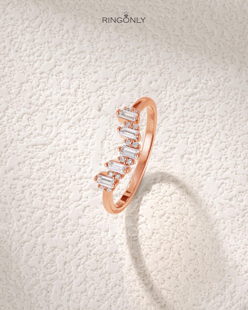 May include: Rose gold ring with a row of baguette-cut and round-cut clear gemstones. The ring has a delicate band and the gemstones are set in a wave-like pattern. The brand name "RINGONLY" is visible in the upper left corner.