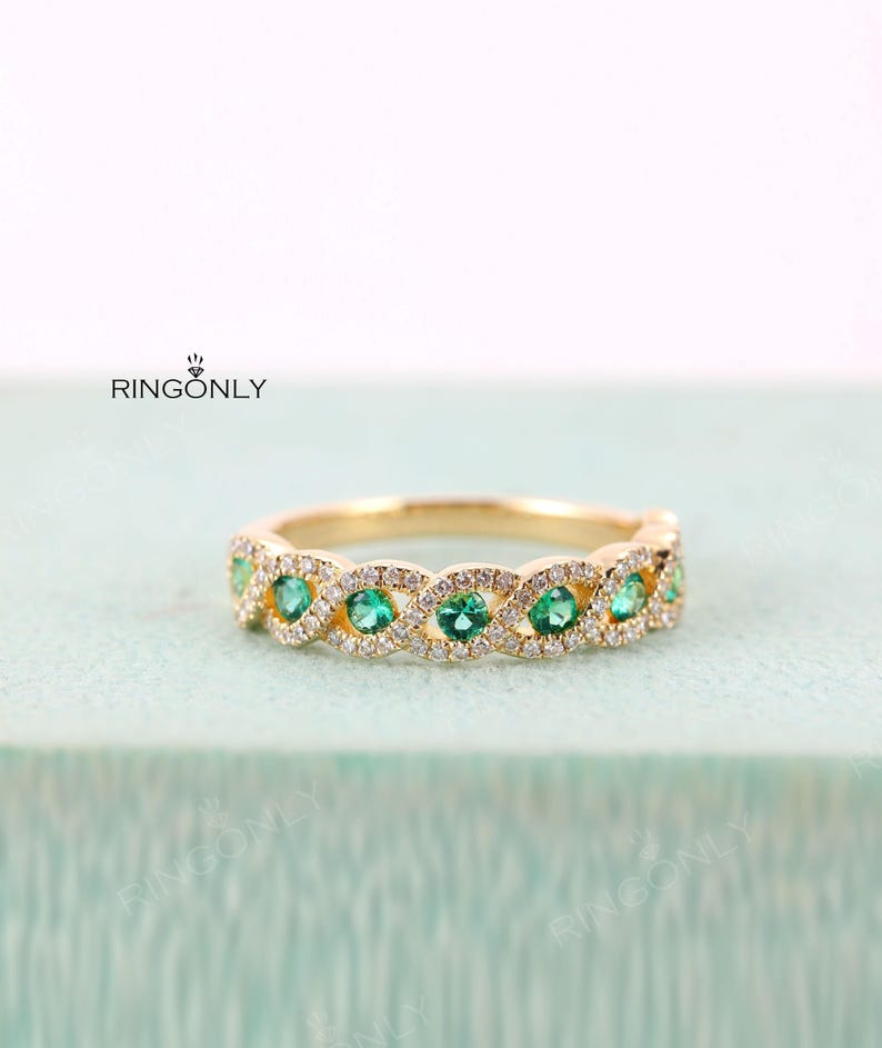 May include: A gold ring with a repeating pattern of emerald green gemstones and small, clear gemstones. The ring's design is interwoven, with the gemstones set in a repeating pattern. The ring is displayed on a light blue surface.