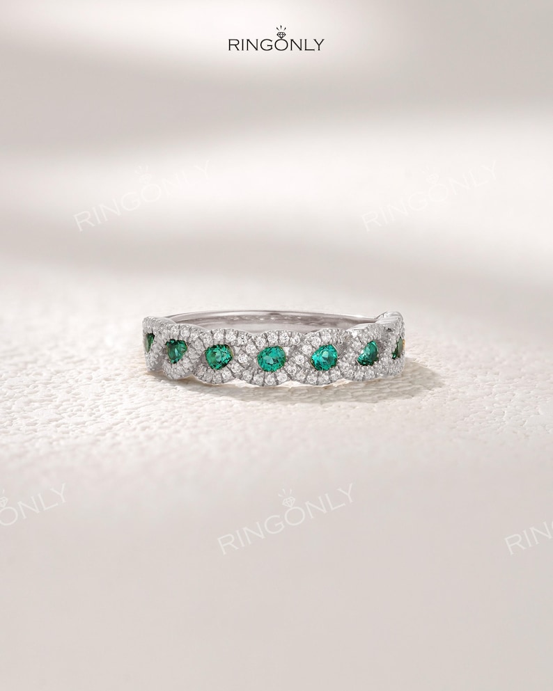 Op de afbeelding: Een zilveren ring met een herhalend patroon van smaragdgroene edelstenen en kleine heldere stenen. De ring is geplaatst tegen een witte achtergrond. De merknaam "RINGONLY" staat bovenaan.