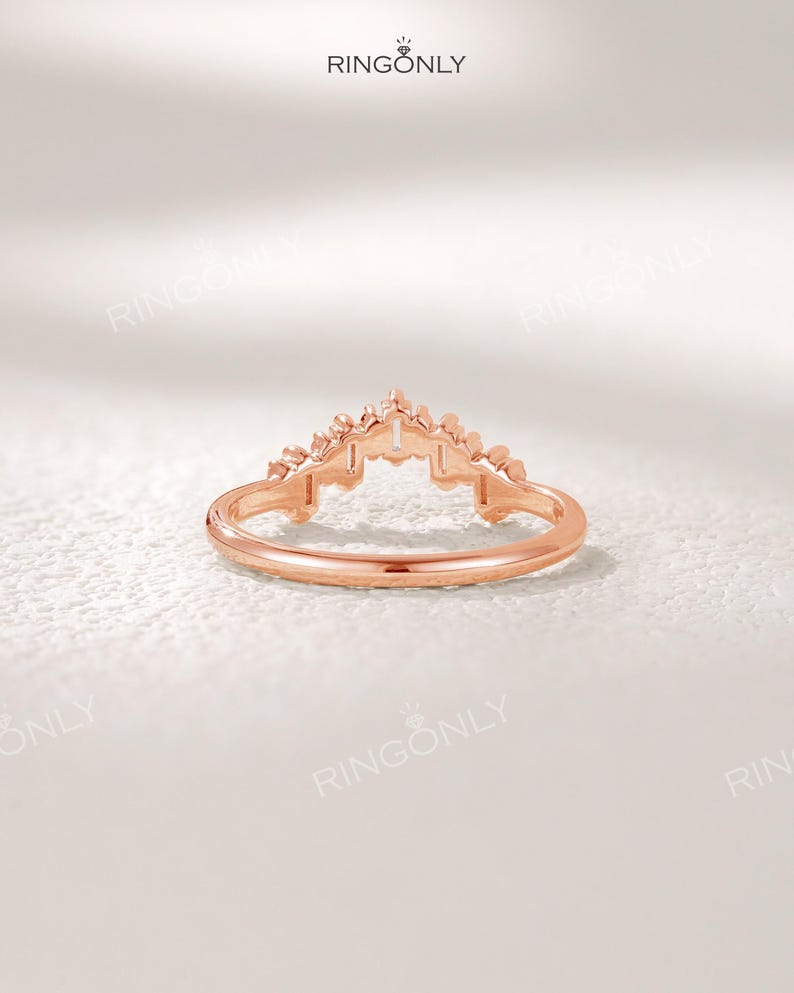 May include: Rose gold ring with a crown-like design. The ring features a delicate band and a detailed, ornate top. The crown design is centered and symmetrical, with small, pointed elements. The ring is set against a soft, white background.