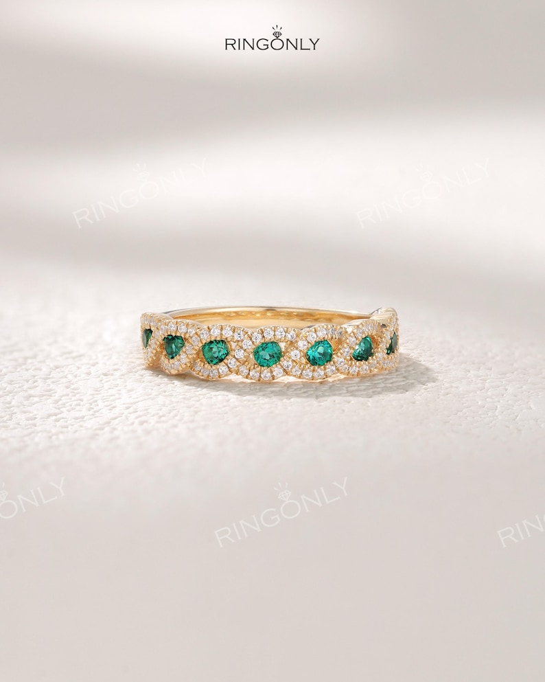 Op de afbeelding: Een gouden ring met een reeks smaragdgroene edelstenen afgewisseld met kleine, heldere edelstenen. De ring heeft een delicaat, sierlijk ontwerp. De merknaam "RINGONLY" is zichtbaar.