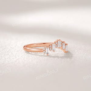May include: Rose gold ring with a curved design. The ring features a row of rectangular, clear gemstones and smaller round gemstones. The brand name "RINGONLY" is visible at the top. The ring is set against a white background.