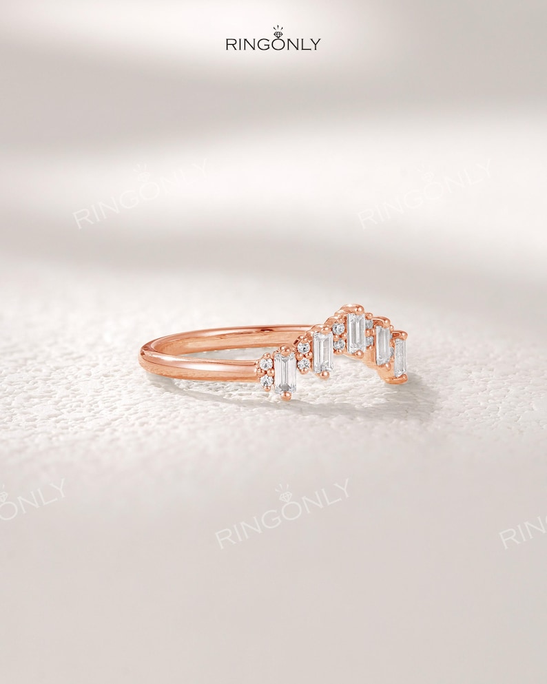 May include: Rose gold ring with a curved design. The ring features a row of rectangular, clear gemstones and smaller round gemstones. The brand name "RINGONLY" is visible at the top. The ring is set against a white background.