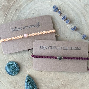 Könnte beinhalten: Zwei Freundschaftsarmbänder mit inspirierenden Botschaften. Das erste Armband ist pfirsichfarben mit einer rosa Steinperle und lautet "Glaube an dich selbst." Das zweite Armband ist burgunderfarben mit einer dunkelgrauen Steinperle und lautet "Genieße die kleinen Dinge."