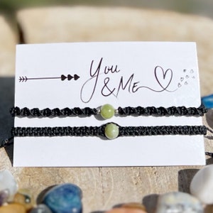 Può includere: Un paio di braccialetti dell'amicizia neri con perline di pietra verde. I braccialetti sono esposti su una carta bianca con il testo "You & Me" e un cuore.