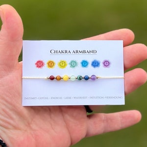 Può includere: Un braccialetto di corda bianca con sette perline colorate che rappresentano i sette chakra. Il braccialetto è su una carta bianca con il testo "CHAKRA ARMBAND" e i sette simboli dei chakra. Il testo sotto i simboli dice "INSTINKT-GEFÜHL-ENERGIE-LIEBE-WAHRHEIT-INTUITION-VERBINDUNG."