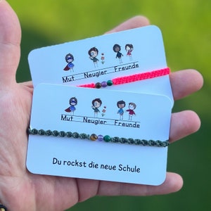 Könnte beinhalten: Zwei weiße Karten mit bunten Illustrationen und dem Text "Mut - Neugier - Freunde" und "Du rockst die neue Schule". Jede Karte hat ein Armband aus Schnur mit bunten Perlen.