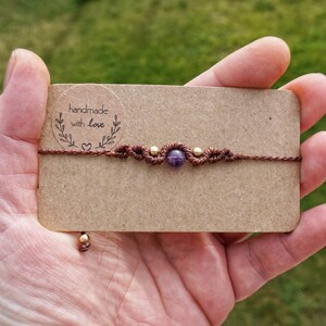 Könnte beinhalten: Ein braunes Kordelarmband mit einem violetten Amethyst-Edelstein und goldenen Perlen. Das Armband befindet sich auf einer braunen Karte mit dem Text "handgefertigt mit Liebe".
