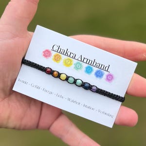 Könnte beinhalten: Ein schwarzes Armband aus Schnur mit sieben bunten Perlen, die die sieben Chakren darstellen. Das Armband liegt auf einer weißen Karte mit dem Text "Chakra Armband" und den sieben Chakra-Symbolen. Der Text unter dem Armband lautet "Instinkt - Gefühl - Energie - Liebe - Wahrheit - Intuition - Verbindung."