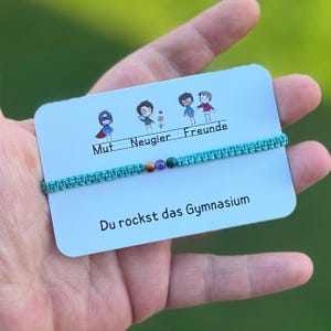 Könnte beinhalten: Ein türkisfarbenes Makramee-Armband mit drei kleinen Perlen wird auf einer Karte mit Cartoonfiguren und deutschem Text präsentiert. Auf der Karte steht "Mut, Neugier, Freunde" und "Du rockst das Gymnasium."