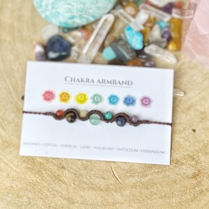 Könnte beinhalten: Ein braunes Chakra-Armband mit sieben bunten Perlen, die die sieben Chakren repräsentieren. Das Armband liegt auf einer weißen Karte mit dem Text "CHAKRA ARMBAND" und "INSTINKT-GEFOHL-ENERGIE-LIEBE-WAHRHEIT-INTUITION-VERBINDUNG".