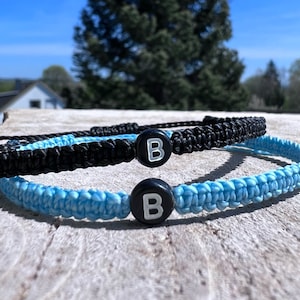 Könnte beinhalten: Zwei Freundschaftsarmbänder mit schwarzen und hellblauen Schnüren, jedes mit einer schwarzen Perle mit einem weißen Buchstaben "B" darauf.