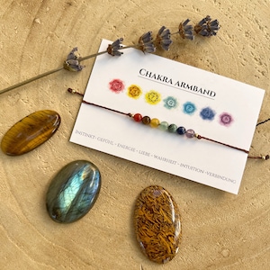 Può includere: Un braccialetto di corda marrone con sette perline colorate che rappresentano i sette chakra. Il braccialetto è su una carta bianca con il testo "Chakra Armband" e i simboli sanscriti di ogni chakra. La carta include anche le parole "Instinkt-Gefühl-Energie-Liebe-Wahrheit-Intuition-Verbindung".