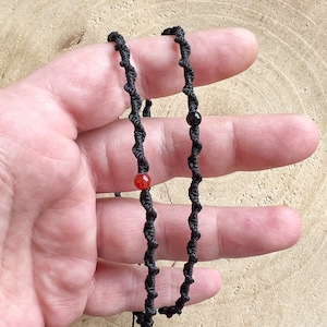 Könnte beinhalten: Schwarze Schnurkette mit roten und schwarzen Perlen. Die Kette ist aus geflochtener Schnur gefertigt und hat ein schlichtes Design.