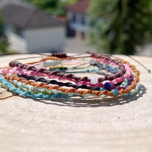 Pu&ograve; includere: Un mucchio di braccialetti intrecciati colorati con piccole perline. I braccialetti sono fatti di diversi colori di corda, tra cui rosa, marrone, blu, verde e arancione. Le perline sono bianche, blu e marroni.