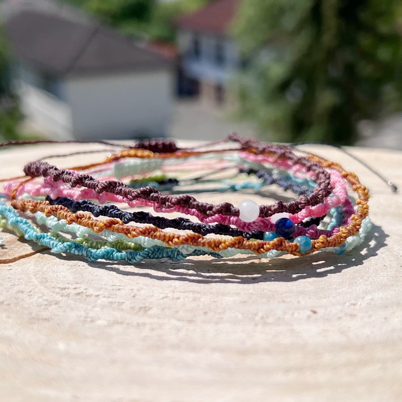 Pu&ograve; includere: Un mucchio di braccialetti intrecciati colorati con piccole perline. I braccialetti sono fatti di diversi colori di corda, tra cui rosa, marrone, blu, verde e arancione. Le perline sono bianche, blu e marroni.