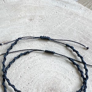 Pu&ograve; includere: Due braccialetti in cordoncino nero con perline argentate, annodati, su uno sfondo in legno chiaro.
