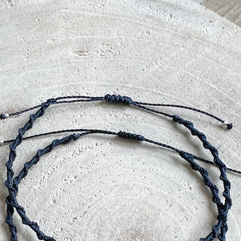 Pu&ograve; includere: Due braccialetti in cordoncino nero con perline argentate, annodati, su uno sfondo in legno chiaro.