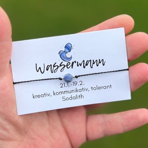 Può includere: Un braccialetto di corda nero con un ciondolo di sodalite blu e un ciondolo d'argento. La carta recita "Wassermann 21.1.-19.2. kreativ, kommunikativ, tolerant Sodalith".