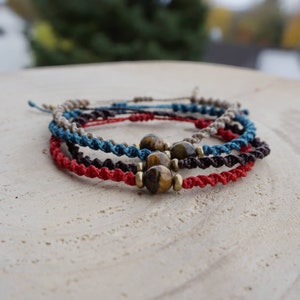 Può includere: Quattro braccialetti intrecciati con un ciondolo a forma di occhio di tigre. I braccialetti sono di colore rosso, marrone, blu e beige.
