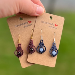 Könnte beinhalten: Zwei Paar handgefertigte Ohrringe mit Edelstein-Akzenten. Ein Paar hat einen dunkelroten Edelstein und das andere Paar einen hellblauen Edelstein. Beide Paare sind aus dunkelblauem und rotem Kordel gefertigt und haben goldene und silberne Ohrhaken. Die Karten, auf denen sie ausgestellt sind, sagen "Handgefertigt mit Liebe".