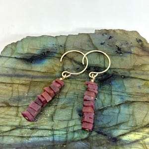 May include: A pair of gold-colored hoop earrings with dangling ruby-colored stone accents. Each earring features a series of small, rectangular stones stacked vertically. The earrings are displayed on a textured, multi-colored stone surface.