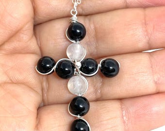 May include: A silver chain necklace with a black and white gemstone cross pendant. The cross is made of wire wrapped around black and white round beads.
