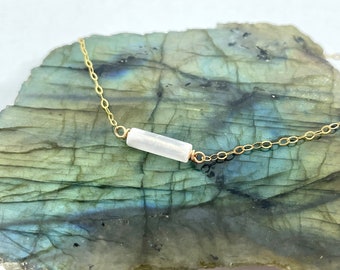 May include: A delicate gold chain necklace with a single white bar pendant. The necklace is displayed on a green and grey stone surface.