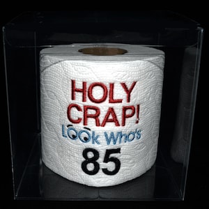 May include: A white toilet paper roll with red and blue embroidery that says "HOLY CRAP! LOOK Who's 85". The roll is in a clear plastic box.