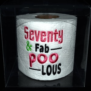 May include: A white toilet paper roll with the text "Seventy & Fab-ulous" embroidered in pink and black. The toilet paper roll is in a clear plastic box.