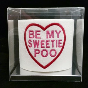May include: A white toilet paper roll with a red heart-shaped patch that says "Be My Sweetie Poo" in pink lettering. The toilet paper roll is in a clear plastic box.