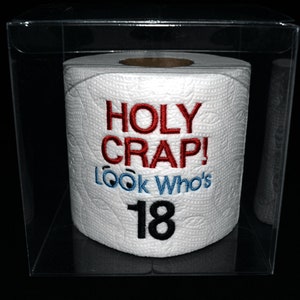 May include: A white toilet paper roll with the text "HOLY CRAP! LOOK Who's 18" embroidered on it in red, blue, and black thread. The roll is in a clear plastic box.