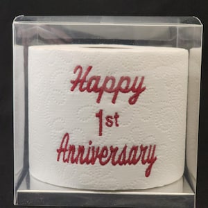 May include: A white toilet paper roll with red embroidery that says "Happy 1st Anniversary". The roll is in a clear plastic box.