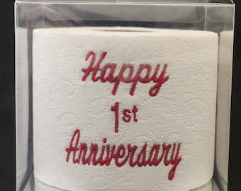 Papel higiénico bordado con la frase "Feliz primer aniversario" en una caja de regalo transparente.