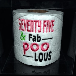 May include: A white toilet paper roll with pink and black embroidery that reads "Seventy Five & Fab-Poo-Lous".