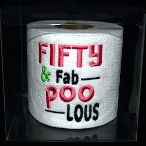 May include: A white toilet paper roll with the text "FIFTY & Fab-POO-LOUS" embroidered on it in pink and black thread. The toilet paper is in a clear plastic box.