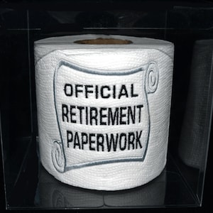 May include: A white toilet paper roll with a gray and black embroidered patch that says "OFFICIAL RETIREMENT PAPERWORK". The roll is in a clear plastic box.